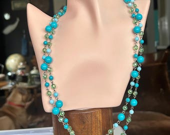 May include: A layered necklace featuring turquoise and light green beads. The necklace is displayed on a mannequin, showcasing the design's two strands of varying bead sizes and colors. The beads are strung on a silver chain.