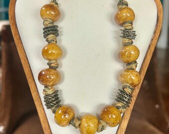 May include: A handcrafted necklace featuring large, round, amber-colored beads interspersed with antique brass-toned metal accents. The necklace is displayed on a wooden jewelry stand, showcasing its unique design and texture.