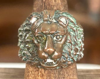 May include: A bronze-colored ring featuring a detailed lion's head. The lion has a textured mane with hints of green patina. The ring is displayed on a wooden stand, showcasing the intricate design and craftsmanship of the jewelry.