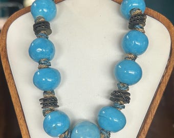 May include: A turquoise beaded necklace with large, round beads. The necklace features dark brown and gold accents between the beads. The necklace is displayed on a white surface, against a wooden stand.
