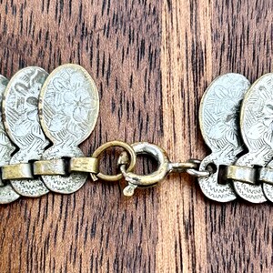 May include: A silver and gold chain necklace with a clasp. The chain is made of small, round links and is decorated with silver coins that have a floral design.