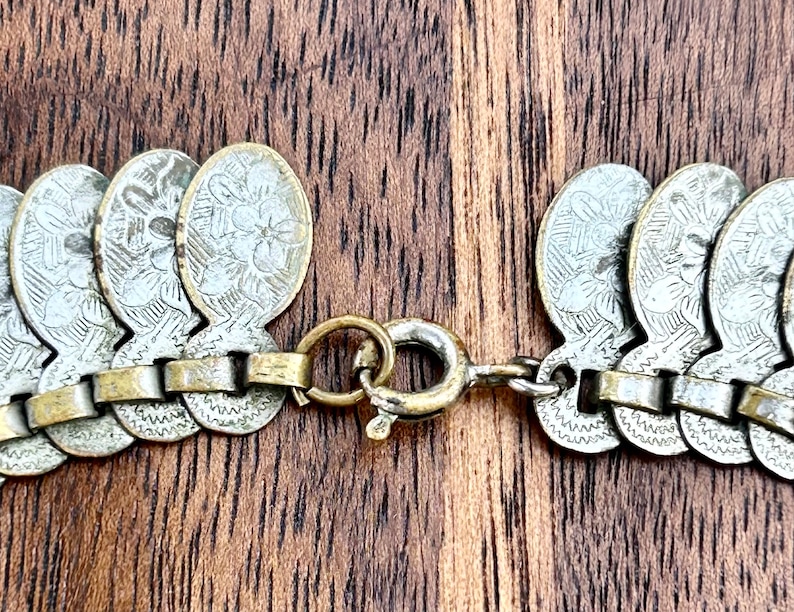 May include: A silver and gold chain necklace with a clasp. The chain is made of small, round links and is decorated with silver coins that have a floral design.