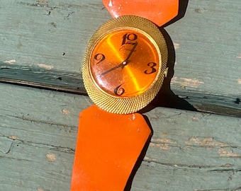 May include: Vintage wristwatch with an orange strap and a gold-toned face. The watch face has a gold-colored border and a gradient orange dial with the numbers 3, 6, 9, and 12. The watch strap is made of orange material.