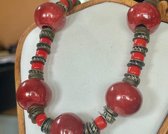 May include: A bold red necklace with large, round beads and smaller red beads. The necklace features antique silver-colored metal accents between the beads. The necklace is displayed on a wooden jewelry stand.