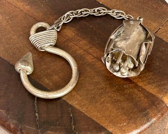 May include: A silver-toned metal keychain featuring a stylized rose pendant and a hook-shaped clasp. The rose pendant has detailed petals and a chain connecting it to the hook. The hook has a pointed end and a wrapped detail near the loop.