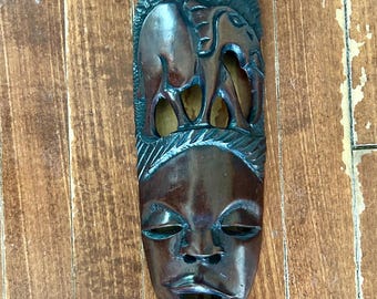 May include: A dark brown, carved wooden mask with a face and two elephants. The mask has a rectangular shape with a face at the bottom and elephants carved at the top. The mask is hanging on a wooden surface.