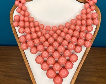 May include: A coral-colored beaded necklace displayed on a white bust with a wooden frame. The necklace features a geometric design of round beads, creating a layered, statement piece. The beads are strung together, forming a V-shape.