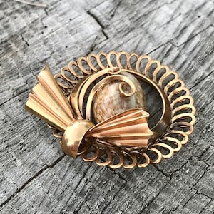 May include: A gold-toned brooch featuring a delicate, intricate design with a shell cameo at the center. The brooch has a bow-like shape with a circular frame.