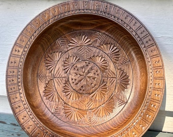 May include: A round, brown wooden bowl featuring intricate hand-carved geometric patterns. The center displays a star-like motif surrounded by radiating fan shapes, while the wide, flat rim is decorated with a repeating triangular border design.