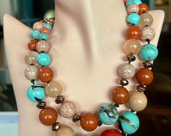 May include: A two-strand necklace featuring large, colorful beads in shades of turquoise, orange, tan, and red. The beads are strung on a black cord and displayed on a mannequin. The necklace has a vintage aesthetic.