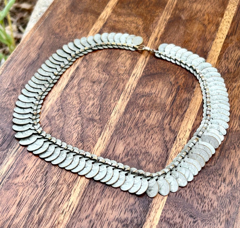 May include: A silver coin necklace with a chain made of small, connected, oval-shaped coins. The necklace is on a wooden surface.