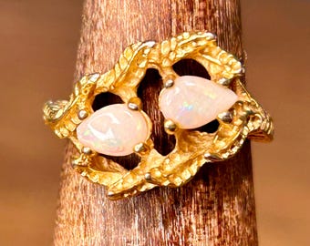 May include: A gold-toned ring featuring two pear-shaped opals. The opals have a milky white color with iridescent flashes. The ring's setting has a textured, organic design, resembling intertwined branches or leaves.