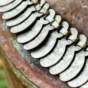 May include: A silver necklace with a decorative design of repeating crescent shapes. The necklace is made of metal and has a vintage look.