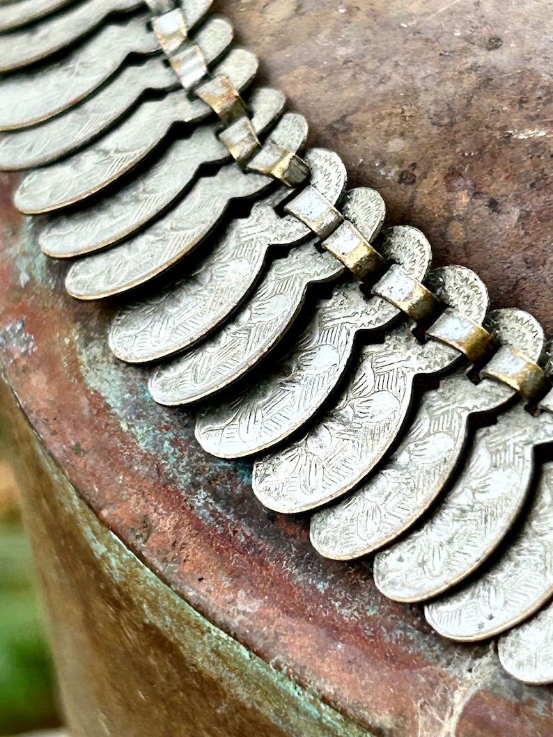 May include: A silver necklace with a decorative design of repeating crescent shapes. The necklace is made of metal and has a vintage look.