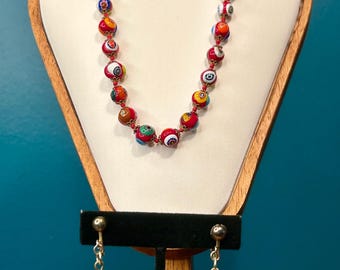 May include: A colorful beaded necklace and matching earrings set. The necklace and earrings feature round, multi-colored beads with intricate patterns. The jewelry is displayed on a wooden stand against a teal background.