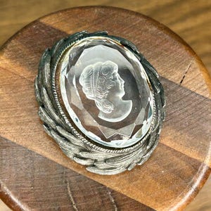 Puede incluir: Broche de camafeo ovalado con una piedra transparente y facetada que representa el perfil de una mujer. La piedra está engastada en un marco de metal plateado con un diseño detallado de hojas. El broche se exhibe sobre una superficie de madera.