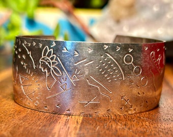 May include: A silver cuff bracelet with etched Native American-inspired designs. The bracelet features a profile of a person wearing a headdress, along with geometric shapes, arrows, and other symbols. The bracelet is displayed on a wooden surface.