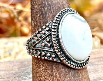May include: A silver-toned ring with an oval, white stone. The ring has a detailed, textured band with small, raised dots and triangular patterns. The stone is set in a decorative bezel.