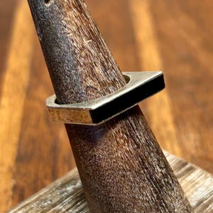 May include: A silver ring with a black rectangular inlay. The ring is displayed on a wooden ring holder.