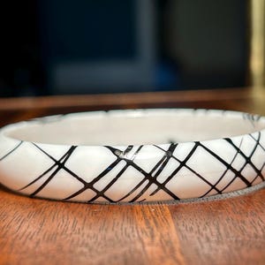 Może przedstawiać: Biała ceramiczna bransoletka z czarnym wzorem krzyżowym. Bransoletka jest okrągła i gładka, z błyszczącym wykończeniem. Czarne linie tworzą geometryczny wzór na powierzchni.
