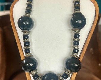 May include: A black beaded necklace with large, round beads and smaller, cylindrical beads. The necklace features silver-toned metal accents and is displayed on a wooden jewelry stand. The necklace is a statement piece.