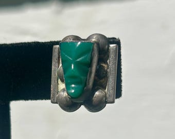 May include: A silver ring featuring a central triangular green stone carved into a stylized face. The metal band is accented with four rounded silver spheres on the sides, framing the green stone in a rectangular setting with smooth edges.