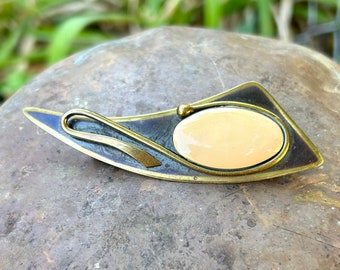 Könnte beinhalten: Eine abstrakte Brosche in Gold und Schwarz mit einem ovalen Cabochon-Stein in blass Pfirsich.