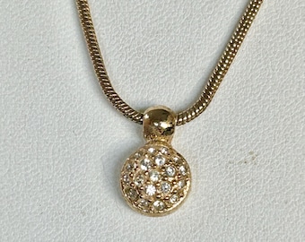 May include: Gold-toned necklace with a round pendant. The pendant is encrusted with small, clear gemstones. The chain has a textured, snake-like design. The necklace is displayed against a neutral background.