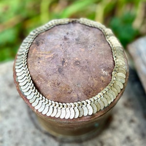 May include: A silver-toned necklace made of linked, coin-shaped charms. The necklace is lying on a brown, textured surface.