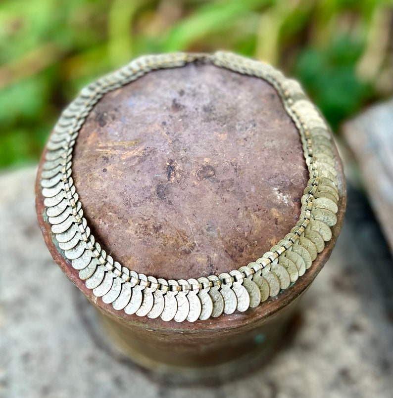 May include: A silver-toned necklace made of linked, coin-shaped charms. The necklace is lying on a brown, textured surface.