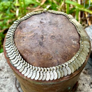 May include: A silver-toned necklace made of linked coin-shaped charms. The necklace is displayed on a brown surface.