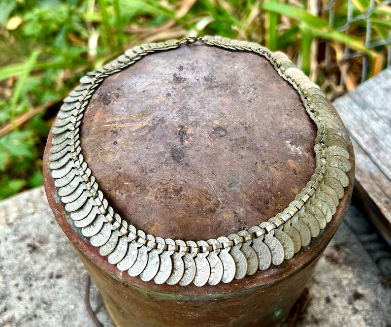 May include: A silver-toned necklace made of linked coin-shaped charms. The necklace is displayed on a brown surface.