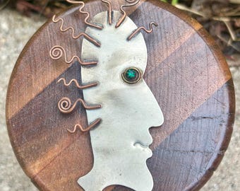 May include: A handcrafted, mixed-media art piece featuring a profile of a face. The face is made of silver metal with a green gemstone eye and copper wire accents. The face is mounted on a round, wooden base with a geometric pattern.