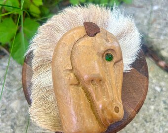 May include: A wooden horse head sculpture with a tan mane and a green gem eye. The horse head is carved from light-colored wood and is mounted on a round, multi-toned wooden base. The mane is made of a light brown and white fur.