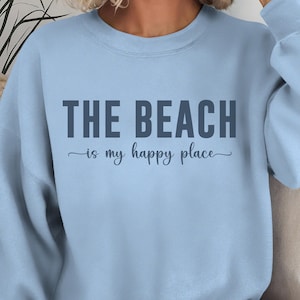 Op de afbeelding: Lichtblauwe trui met de tekst "THE BEACH is my happy place" in donkerblauw.
