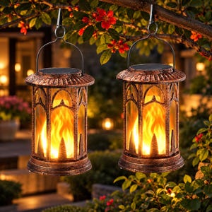 May include: Two hanging solar lanterns with a distressed bronze-colored metal frame and clear glass panels. Each lantern features a top-mounted solar panel, a metal handle, and an internal light source that mimics the appearance of flickering flames.
