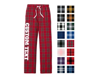 May include: Red plaid pajama pants featuring a white drawstring waist and the text CUSTOM TEXT printed vertically in white on the right leg. A grid of eighteen additional plaid color pattern swatches is displayed to the right of the pants.