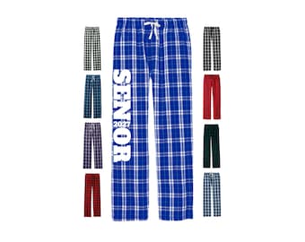 May include: Blue plaid pajama pants with a white drawstring waist and the vertical white text SENIOR 2027 printed on the left leg. The image also displays eight additional pairs of plaid pajama pants in various color combinations.