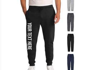 May include: Black sweatpants with white text reading "YOUR TEXT HERE" down the left leg. The pants have a drawstring waist and are shown with white sneakers. Additional colors of gray, charcoal, black, and navy are displayed.