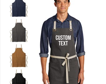 May include: A selection of aprons in navy, charcoal, brown, and black. The aprons have a pocket and adjustable neck strap. The main apron in the image is dark gray with the text "CUSTOM TEXT" in white.