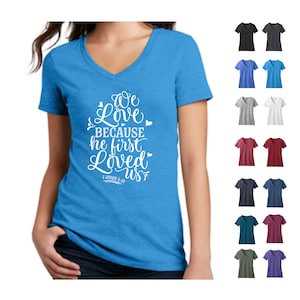 May include: A light blue v-neck t-shirt with the text "We love because he first loved us" and the scripture reference "1 John 4:19". The shirt is shown on a model and is available in a variety of colors.