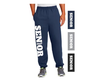 May include: Navy blue sweatpants featuring the word SENIOR printed vertically in large white capital letters along the right leg, with the year 2027 positioned between the letters N and I. The design is also shown in three color variations.