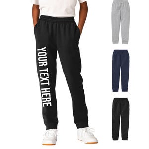 May include: Black sweatpants with the text "YOUR TEXT HERE" in white down the left leg. The image also shows gray, navy blue, and black sweatpants. The person is wearing white sneakers and a white t-shirt.