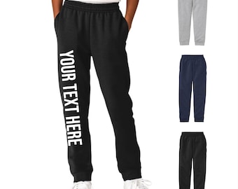 May include: Black sweatpants with the text "YOUR TEXT HERE" in white down the left leg. The image also shows gray, navy blue, and black sweatpants. The person is wearing white sneakers and a white t-shirt.