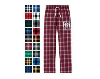 May include: Maroon plaid pajama pants with a white drawstring waistband and the text YOUR TEXT HERE printed in white on the upper thigh. A vertical color palette of twenty different plaid fabric swatches is displayed to the left of the pants.