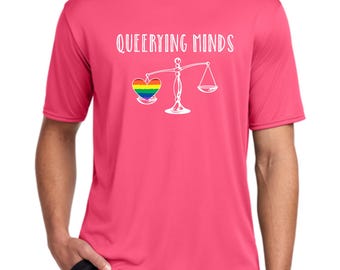 May include: A short-sleeved, pink t-shirt with the text "Queerying Minds" above a graphic of a rainbow heart on a scale. The scale is white. The shirt is made of a lightweight material.