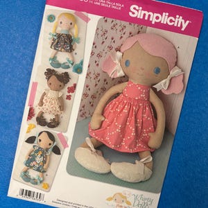 May include: A Simplicity sewing pattern for Whimsy Dolls, featuring a large doll with a pink dress and smaller doll illustrations. The pattern includes the text "U.S. ONE SIZE" and "SINCE 1927". The main doll has a pink head and white shoes.
