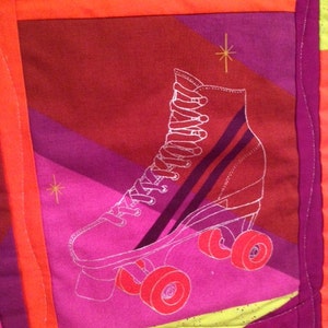 May include: A colorful quilt featuring a retro roller skate design. The skate is outlined in white on a pink background, with red wheels and a purple stripe. The quilt has a geometric pattern with orange, red, and purple blocks.