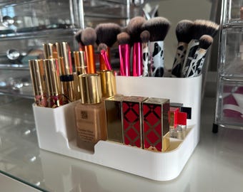 Puede incluir: Un organizador de maquillaje de plástico blanco lleno de varios productos de belleza. El organizador contiene brochas de maquillaje, barras de labios y base de maquillaje. Las brochas tienen mangos con diseños en blanco y negro y rosas. Las barras de labios tienen envases dorados.