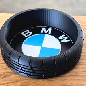 Puede incluir: Una bandeja circular negra con un diseño de banda de rodadura de neumático alrededor del borde. El centro presenta el logotipo de BMW en blanco y azul. La bandeja probablemente está hecha de plástico y se encuentra sobre una superficie de madera.